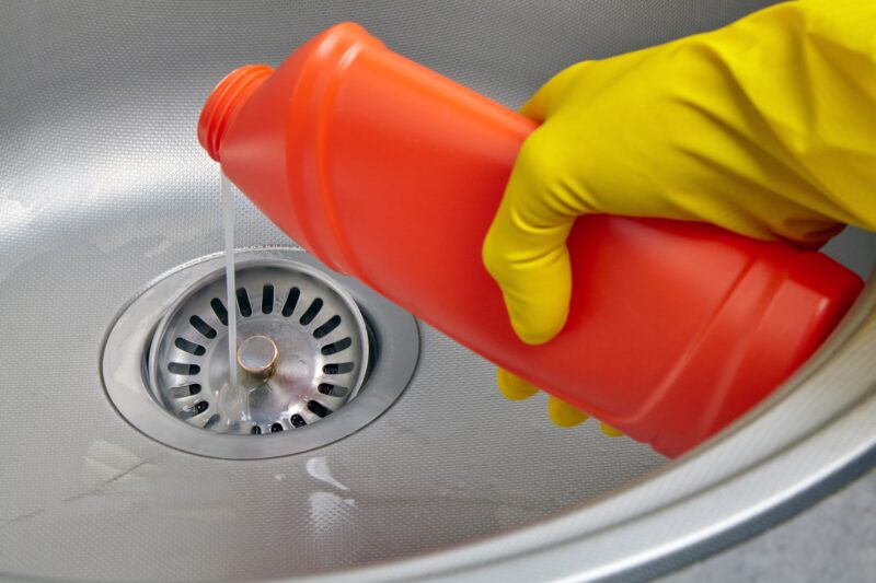 homeowner wearing a yellow rubber glove while pouring a bottle of drain cleaning solution down kitchen sink drain